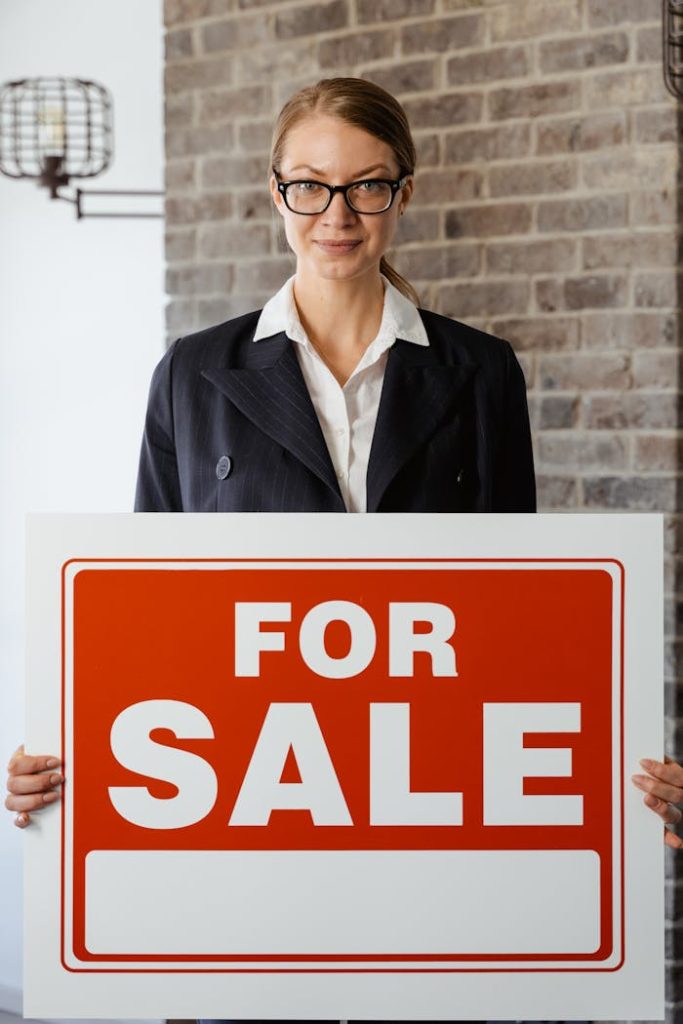 Confident woman in corporate attire holding a for sale sign against a brick wall.
