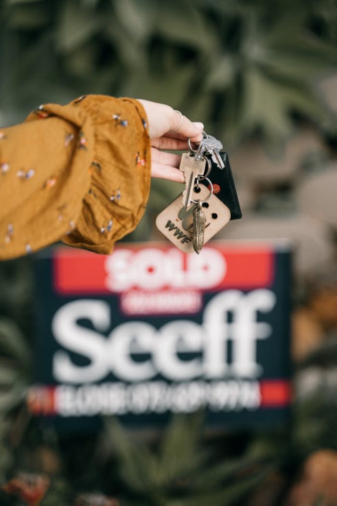 A hand holds keys in front of a blurred sold real estate sign, symbolizing completed sale.