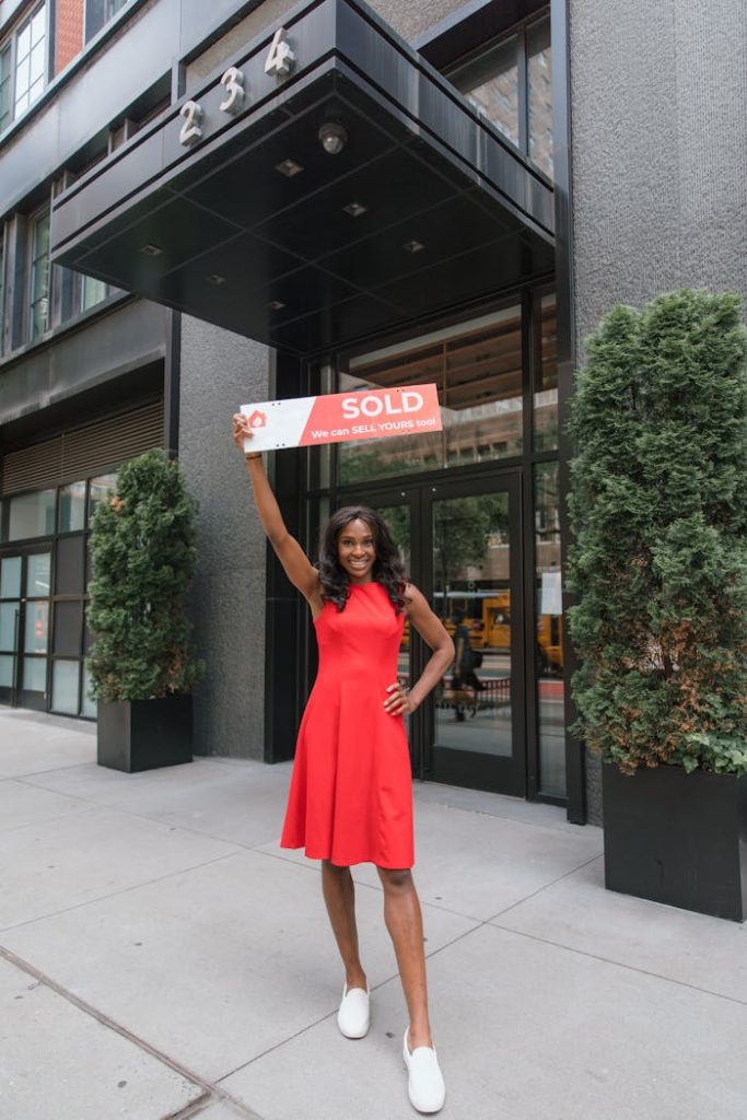 Smiling woman in red dress holding a sold sign outside a building, showcasing real estate success.